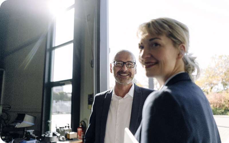 A female professional gazing confidently towards the camera, with a male colleague in the background, set within an office environment.