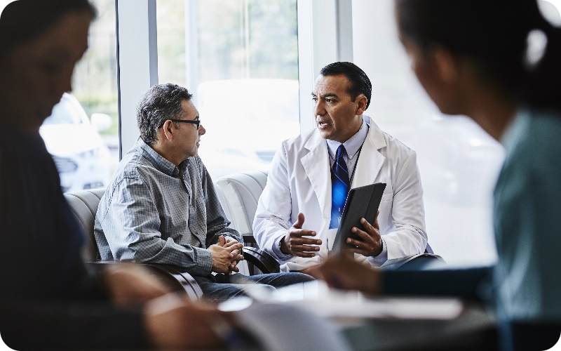 A healthcare professional seated beside a man, holding an iPad and providing an informative explanation.