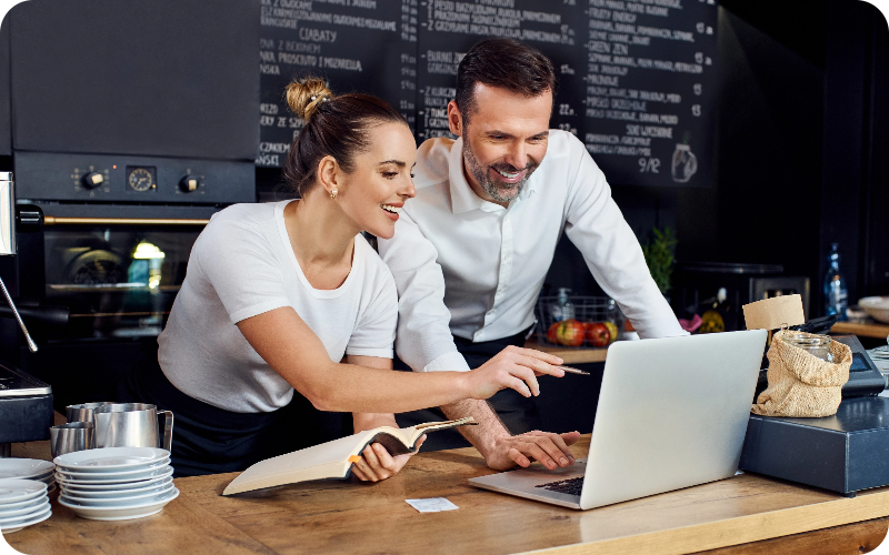 A woman and a man positioned behind a café counter, leaning over a laptop and gesturing towards the screen in a focused manner.