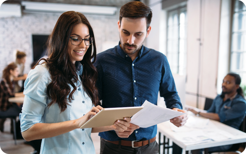 An office scene featuring a woman holding an iPad and a man holding papers. The woman is engaged in explaining something to the man, fostering a collaborative environment.